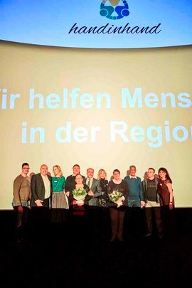 eine Reihe von Menschen, 2 davon mit einem Blumenstrauß stehen vor dem Logo "Wir helfen Menschen in der Region